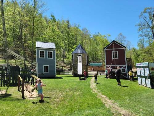 Children play in a grassy area with colorful small houses and trees in the background on a sunny day.