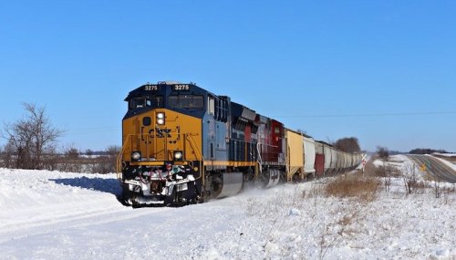 A freight train travels through a snowy landscape under a clear blue sky.