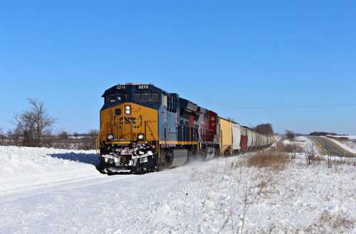 A freight train travels through a snowy landscape under a clear blue sky, with a road visible in the background.