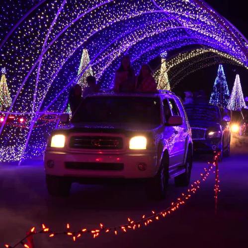 A car drives through a colorful light tunnel with festive decorations and illuminated trees in the background.