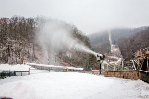 Snowmaking machine spraying artificial snow on a winter slope, surrounded by trees and a snowy landscape.
