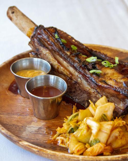 A large beef rib on a wooden plate, accompanied by two dipping sauces and a side of kimchi.