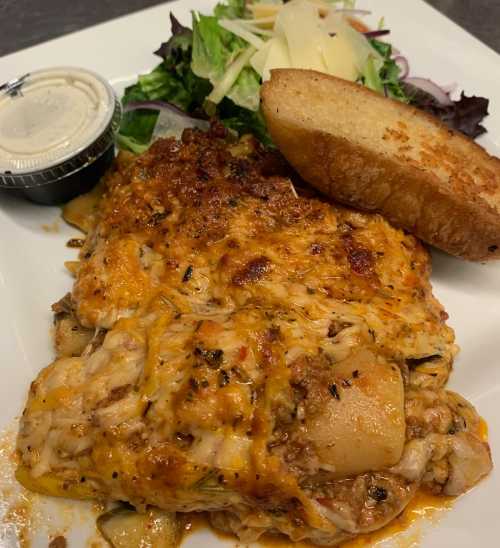 A plate of cheesy lasagna with a side salad and garlic bread, served with a small container of dressing.