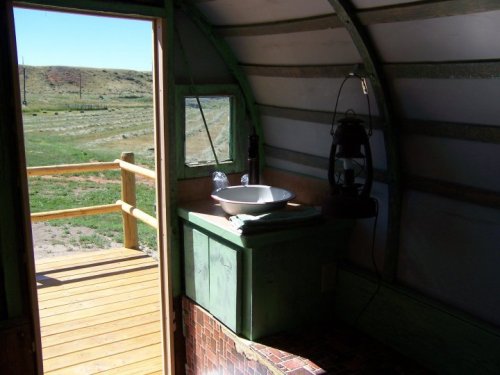 Interior of a rustic cabin with a sink, lantern, and a view of a grassy landscape through an open door.