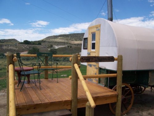 A rustic wagon on a wooden deck, surrounded by open fields and blue skies, with a sign reading "Sheep Wagon."