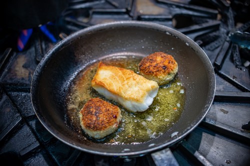 A skillet on a stove with a piece of fried fish and two golden-brown fish cakes in sizzling oil.