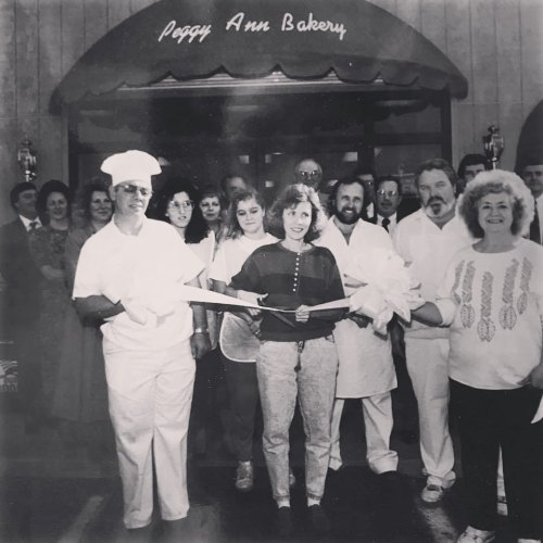 A group of people, including bakers, gather to celebrate the opening of Peggy Ann Bakery, holding a ribbon for a ceremony.
