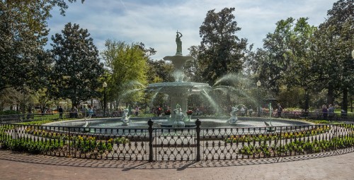 A scenic fountain surrounded by greenery and flowers, with a statue at the center and people enjoying the park.