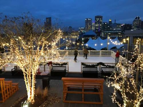 A winter scene featuring an ice skating rink surrounded by twinkling lights and a city skyline at dusk.