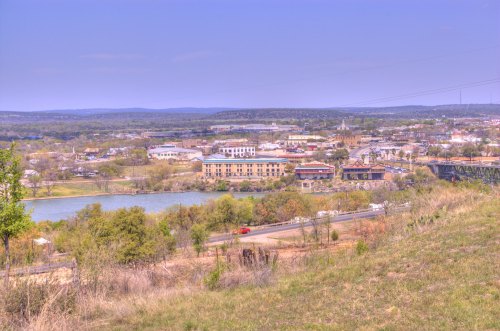 A scenic view of a town by a river, with greenery in the foreground and distant hills under a clear blue sky.