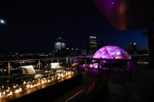 A rooftop terrace at night featuring a glowing dome, fire pit, and city skyline in the background.