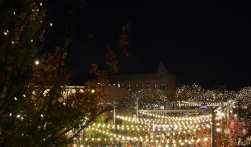 A nighttime scene filled with twinkling lights strung across a festive outdoor area, surrounded by trees and buildings.