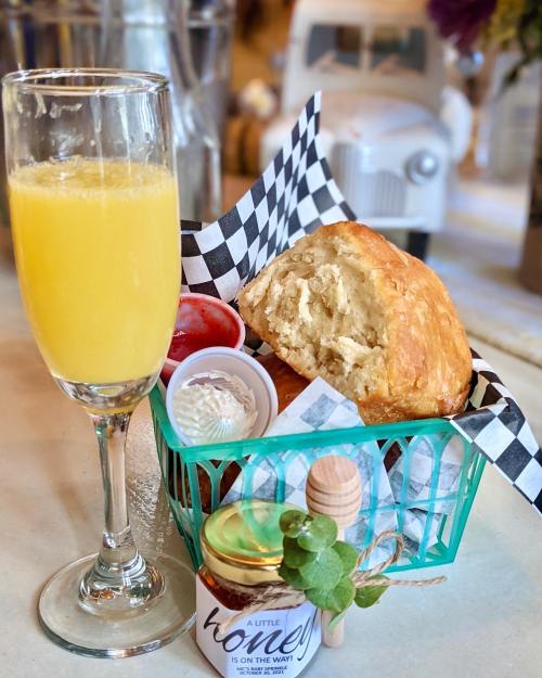 A glass of mimosa beside a basket of bread rolls, honey, and condiments on a table.