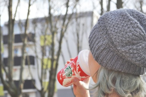 A person with light blue hair wearing a gray beanie drinks from a festive Starbucks cup, surrounded by trees.