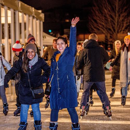 Two women smile and skate on an ice rink at night, surrounded by other skaters and festive lights.