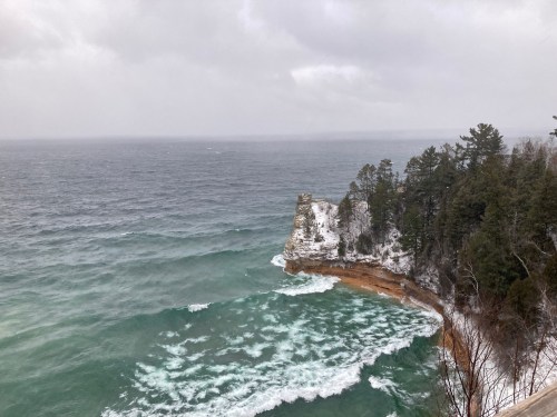 A rocky coastline with waves crashing against the shore, surrounded by snow-covered trees under a cloudy sky.