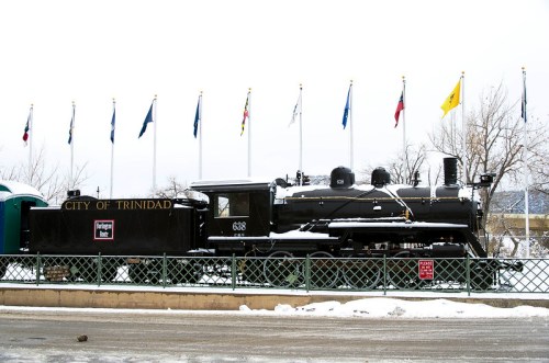 A vintage black steam locomotive named "City of Trinidad" displayed outdoors, surrounded by snow and flags.