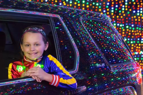 A smiling child in a colorful jacket leans out of a car window, surrounded by vibrant holiday lights.