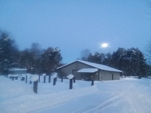 A snowy landscape at dusk featuring a house and trees, with a bright moon visible in the sky.