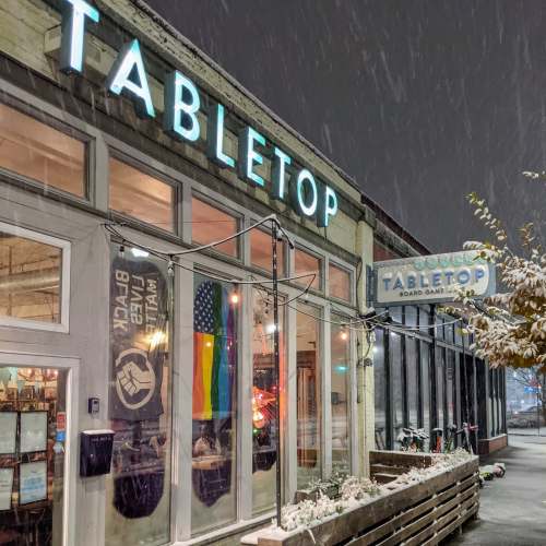 A cozy board game café named "Tabletop," with snow falling outside and colorful signage.