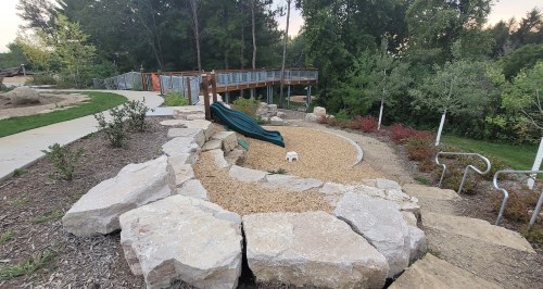 A playground area with a slide, surrounded by rocks and gravel, set in a green park with trees in the background.