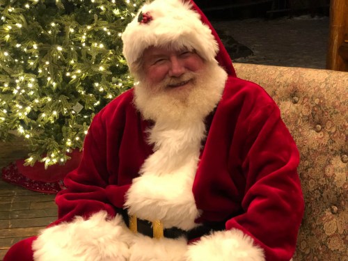 A jolly Santa Claus in a red suit with white fur, sitting on a chair in front of a decorated Christmas tree.