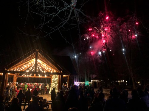 A festive outdoor scene at night with a crowd watching fireworks above a decorated pavilion.