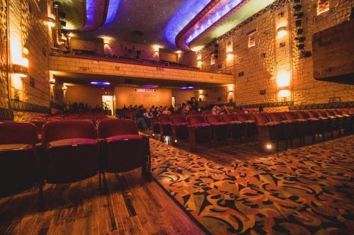Interior of a vintage theater with empty red seats, patterned carpet, and dim lighting, showing a few audience members.