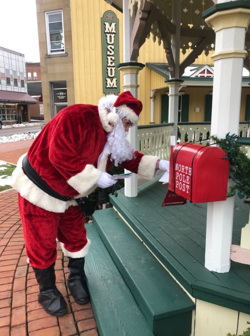 Santa Claus in a red suit placing a letter in a North Pole post mailbox outside a museum. Snow on the ground.