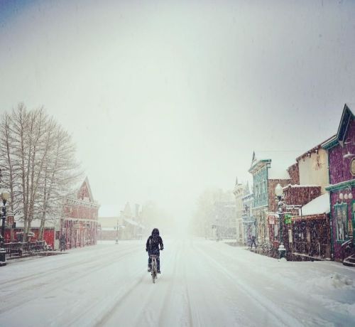 A person rides a bicycle down a snowy street lined with colorful buildings, surrounded by a wintery landscape.