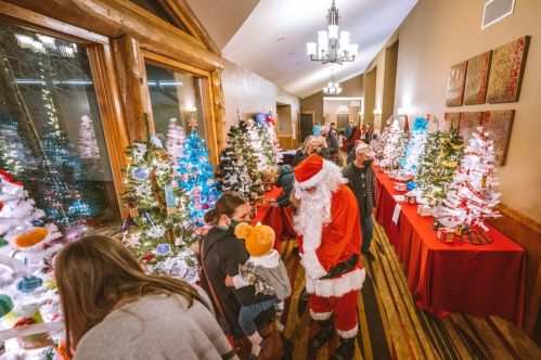 A festive scene with decorated Christmas trees, families, and Santa interacting in a warmly lit hall.