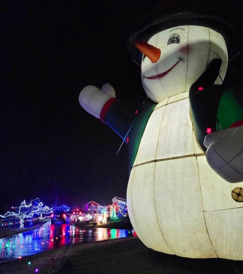 A large, illuminated snowman with a cheerful expression stands in a festive, decorated outdoor setting at night.