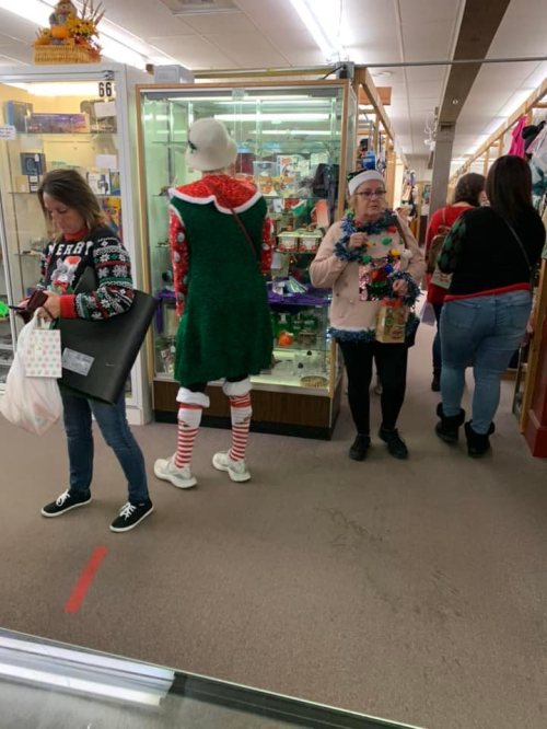 A festive scene in a store with shoppers, including a person dressed as an elf in a green outfit and striped stockings.