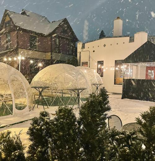 Snow falls gently on a winter scene featuring transparent igloo-like structures and a cozy building in the background.
