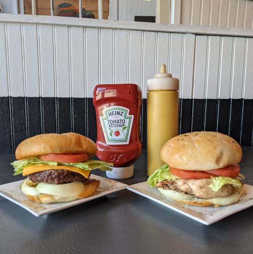 Two burgers on plates, one beef and one chicken, with lettuce, tomato, and condiments of ketchup and mustard in the background.