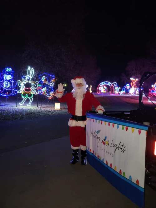 A person dressed as Santa waves in front of colorful holiday lights at a festive nighttime display.