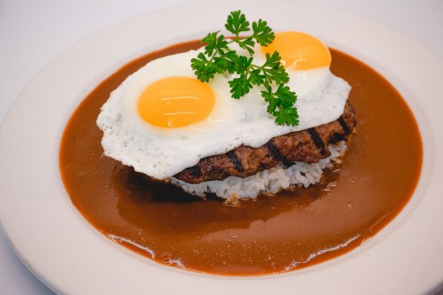 A plate of rice topped with a grilled patty, two fried eggs, and a rich brown sauce, garnished with parsley.