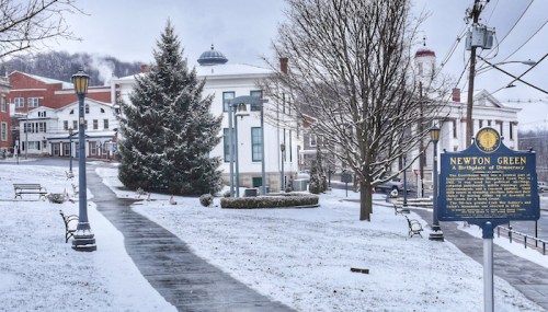 Snow-covered park with benches, a Christmas tree, and a historical marker, surrounded by buildings in a small town.