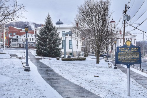 Snow-covered park with a path, benches, a Christmas tree, and a historical sign in a small town.