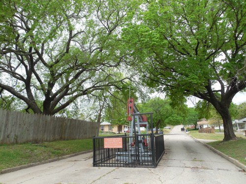 A pump jack sits on a residential street, surrounded by green trees and a wooden fence.