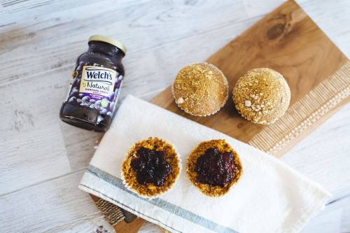 A wooden board with three muffins, two halved with grape jelly, and a jar of Welch's grape jam beside them.