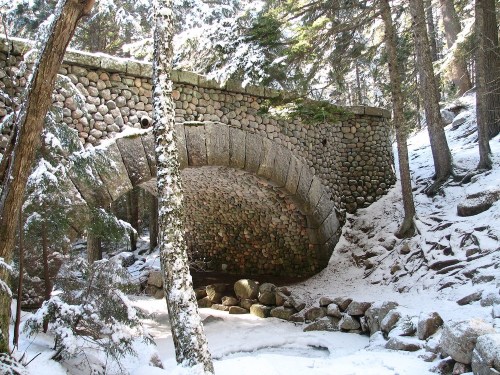 A stone arch bridge in a snowy forest, surrounded by trees and rocky terrain.