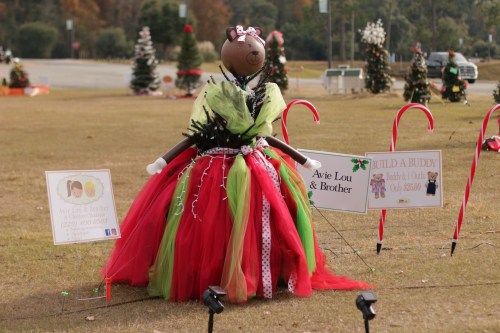 A festive scarecrow dressed in a colorful tutu, surrounded by candy canes and holiday decorations in a park setting.