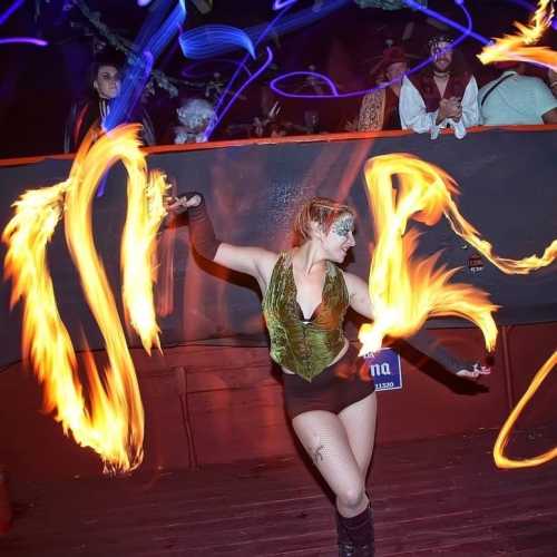 A performer twirls flaming poi in a vibrant, colorful setting, surrounded by an audience enjoying the show.