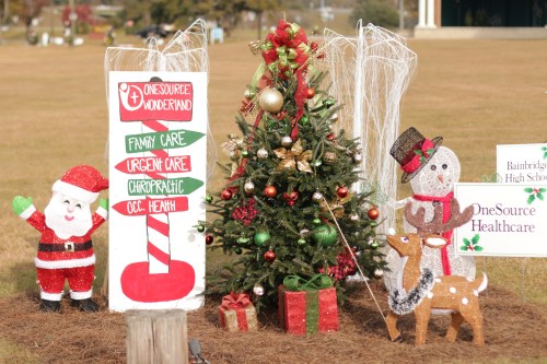 A festive scene featuring a decorated Christmas tree, Santa, a snowman, and holiday signs in a grassy area.