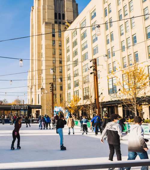 A bustling outdoor ice skating rink surrounded by tall buildings and autumn trees, with people enjoying the activity.