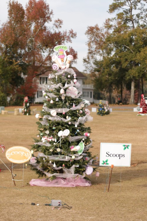 A decorated Christmas tree with pink accents and a sign reading "Scoops" in a grassy area with other trees in the background.