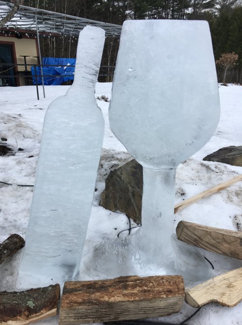 An ice sculpture of a wine bottle and a large wine glass, set on snowy ground with wooden logs nearby.