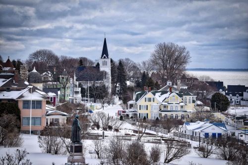 A snowy coastal town with colorful houses, a church steeple, and a statue in the foreground under a cloudy sky.