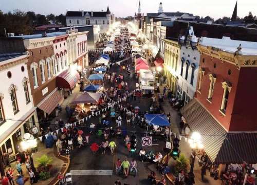 Aerial view of a bustling street festival with tents, crowds, and historic buildings under a twilight sky.